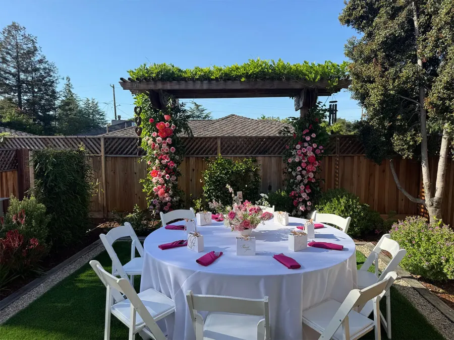 Pink and red rose arch framing a round garden table with white linens and fuchsia napkins — floral installation styling for Oakland catering events